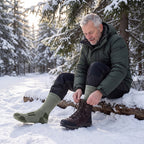 Man i armégröna Arctic Thermal Socks knyter kängorna på en stock i vinterskogen